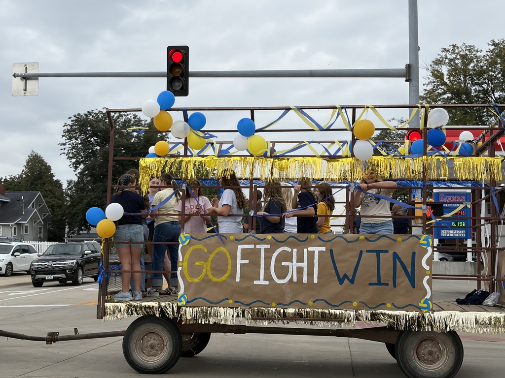 Students ride a blue-and-gold parade float decorated with balloons and a large “GO FIGHT WIN” sign.