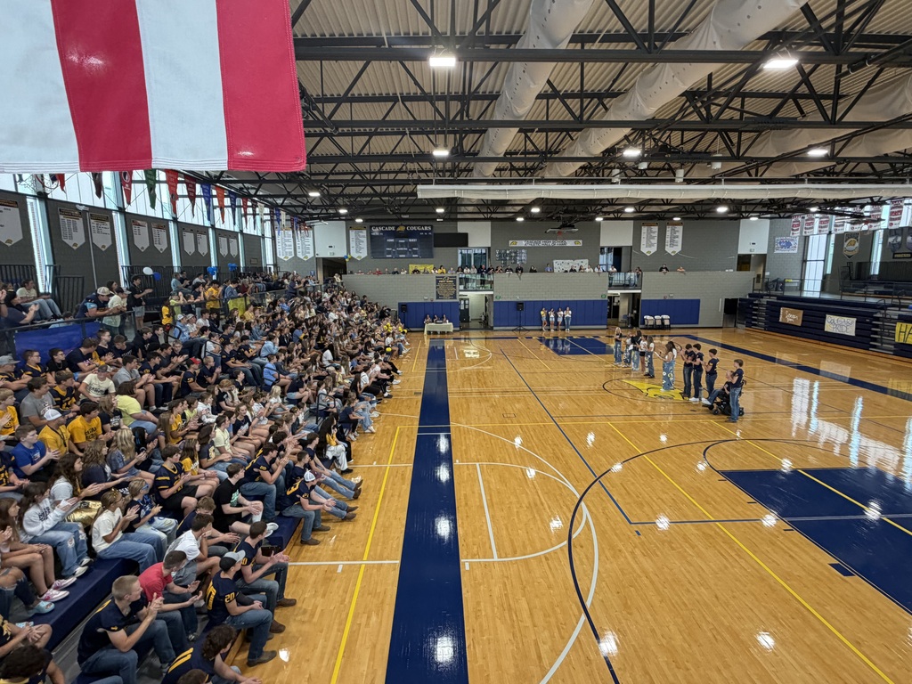 A packed gym of students applauds from the bleachers while classmates stand at center court during a pep rally.