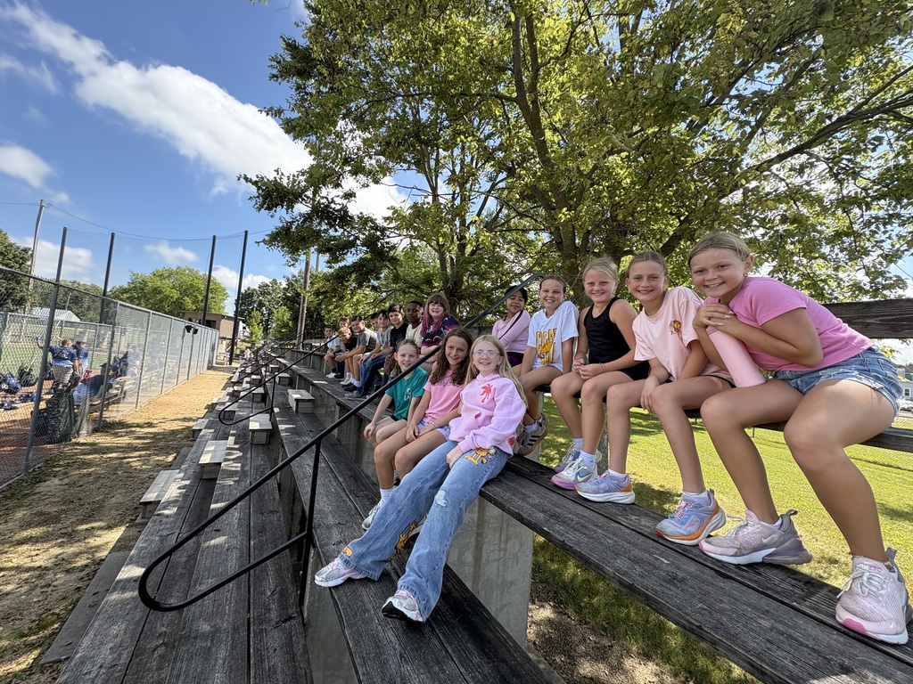 Students sit together on wooden bleachers beside a baseball field, smiling for a group photo under leafy trees.