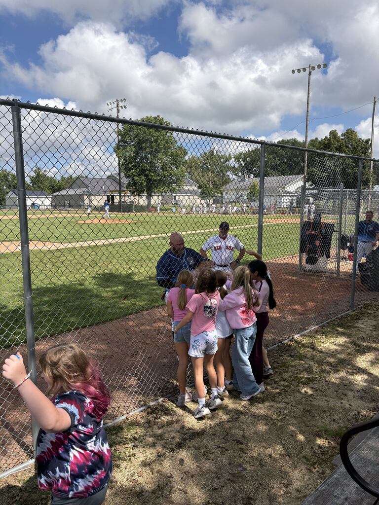 Students gather along a chain-link fence to greet baseball players during a sunny afternoon at the ballpark.