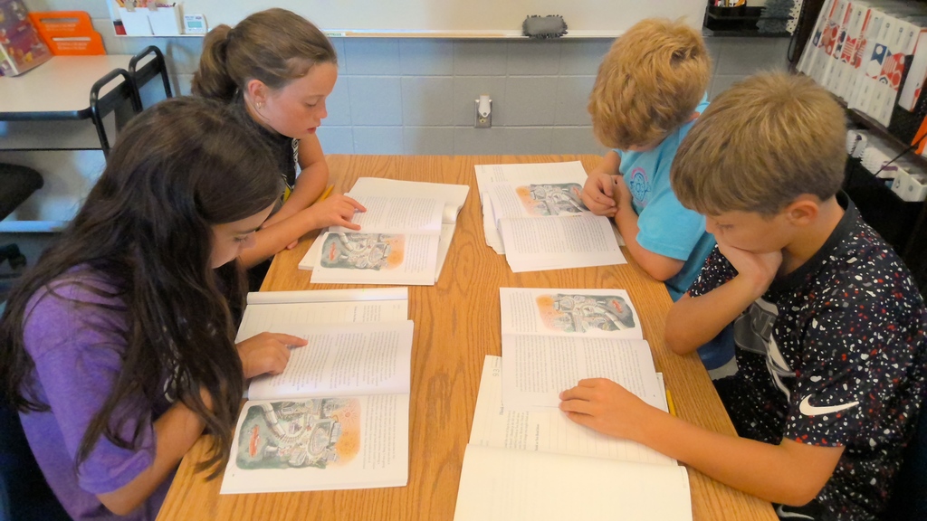 Four students sit around a classroom table, focused on reading illustrated books and completing worksheets.