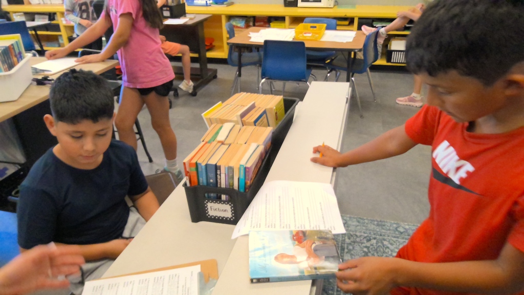 Students work at classroom stations with books and worksheets, including a basket labeled “Fiction.”