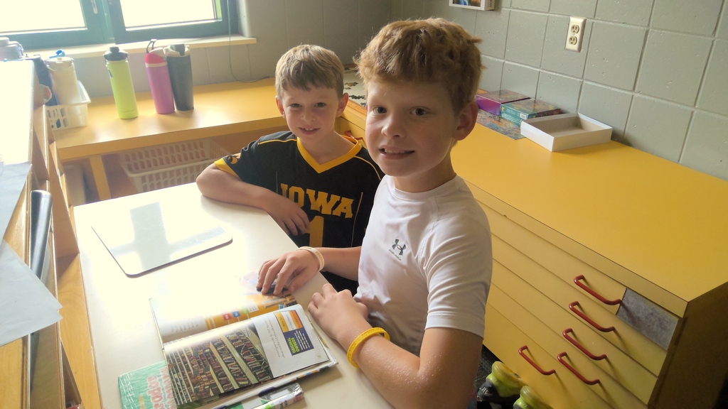 Two students smile at a classroom table with open books and school supplies in a bright reading area.