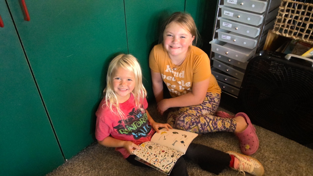 Two students sit on the classroom floor smiling together, with one holding an open picture book.