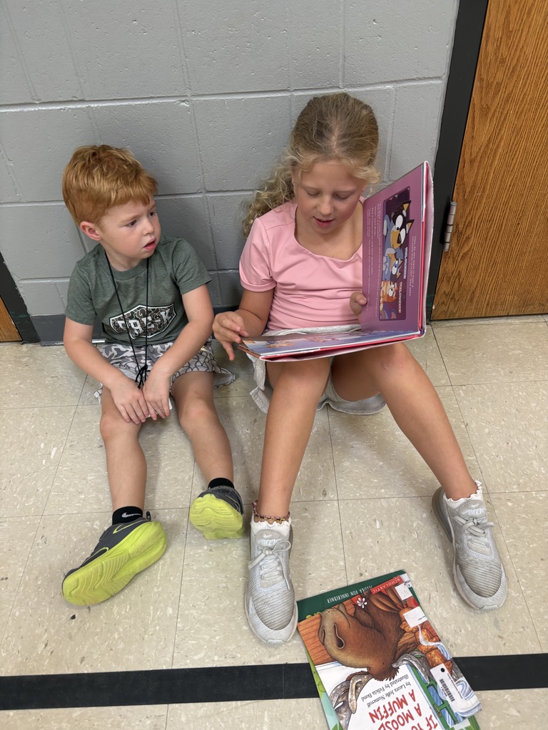 An older child reads a picture book to a younger child as they sit together on the floor against a school hallway wall.