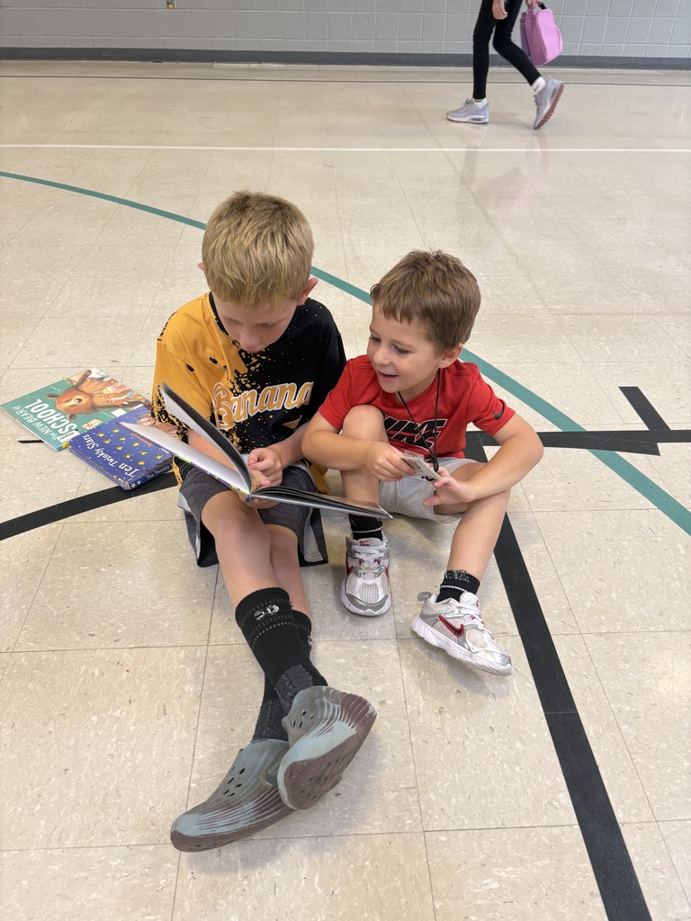 Two children sit together on a gym floor, looking through an open picture book with additional books beside them.