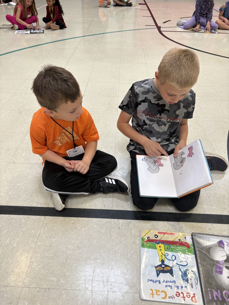 Two children sit cross-legged on a gym floor as one points to an open picture book; other students read in the background.