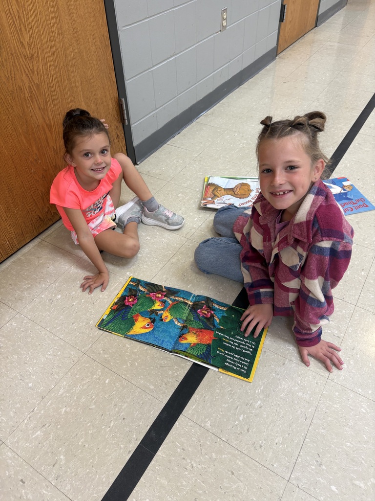 Two smiling children sit beside an open, colorful picture book on the floor near a school hallway door.