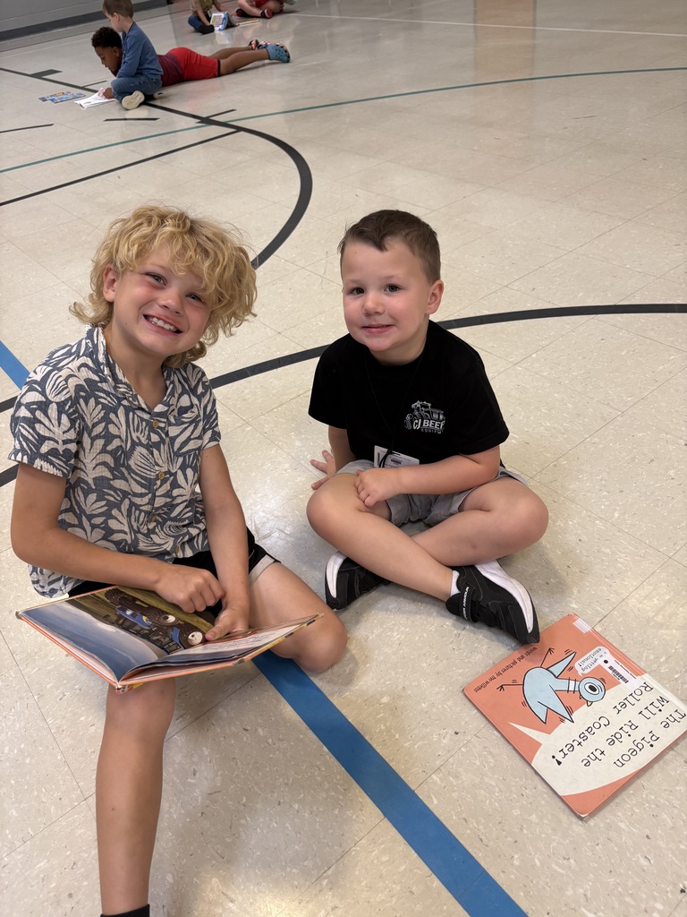 Two smiling children sit cross-legged on a gym floor with open picture books while other students read in the background.