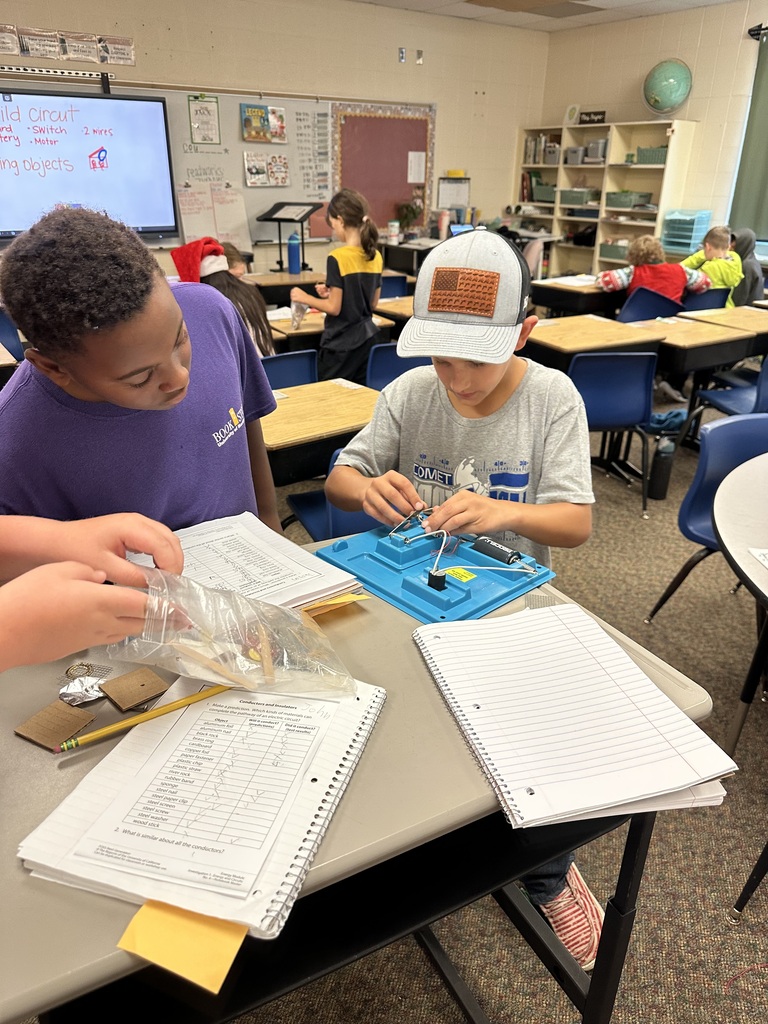 Students collaborate at a classroom table to build and test a simple electrical circuit using wires, a battery, and other components.