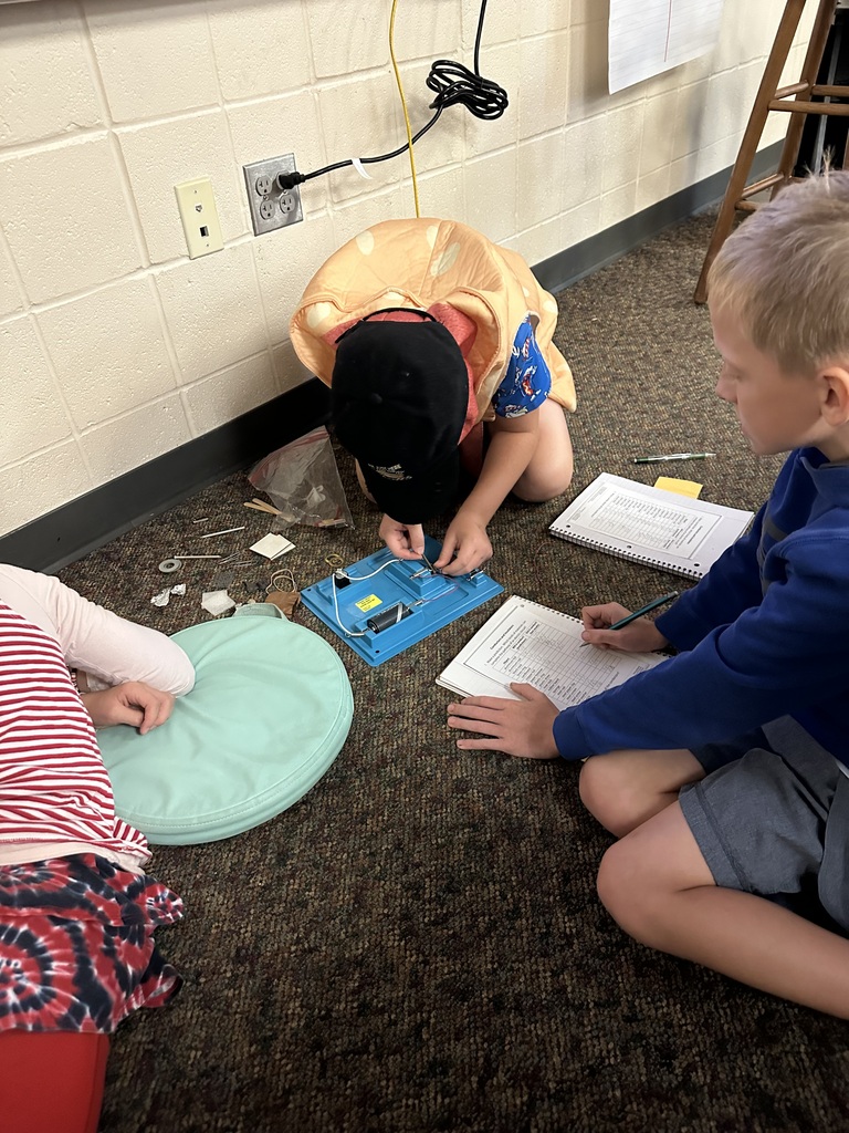 Three students work together on the floor, assembling a simple electrical circuit while recording observations on a worksheet.