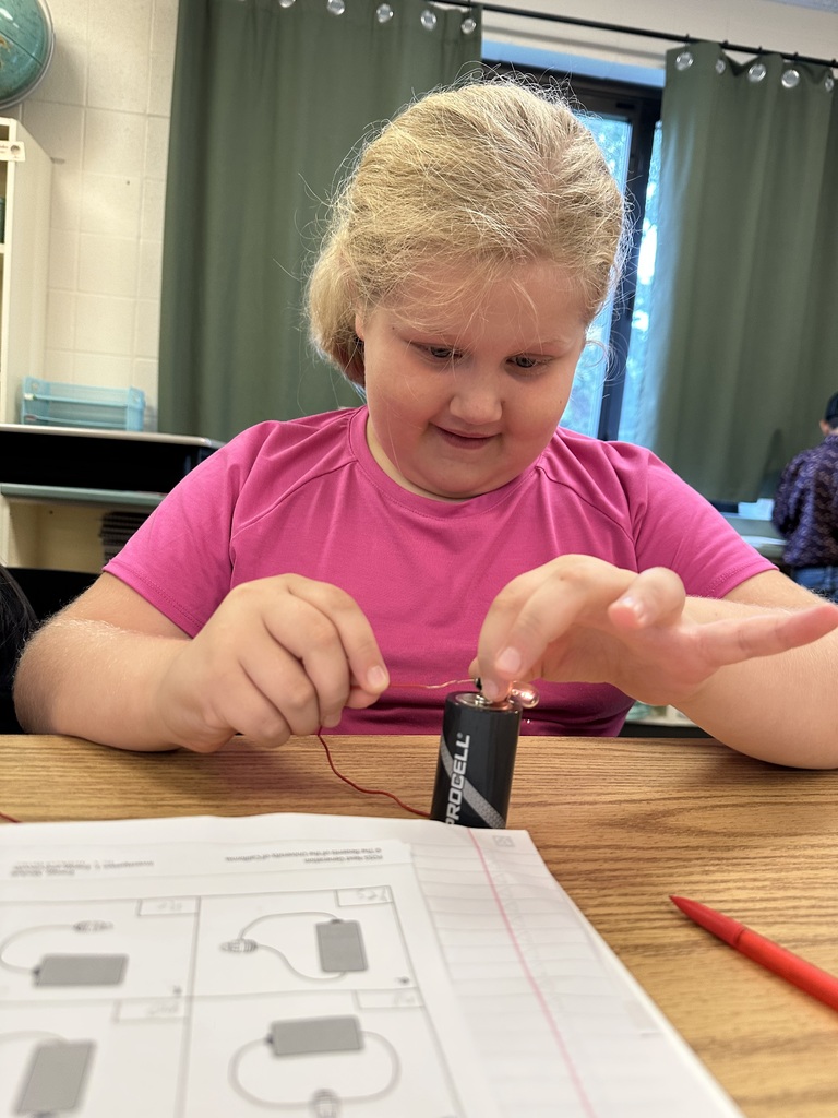 A student in a pink shirt concentrates on connecting a wire to a battery during a classroom electrical circuit activity.