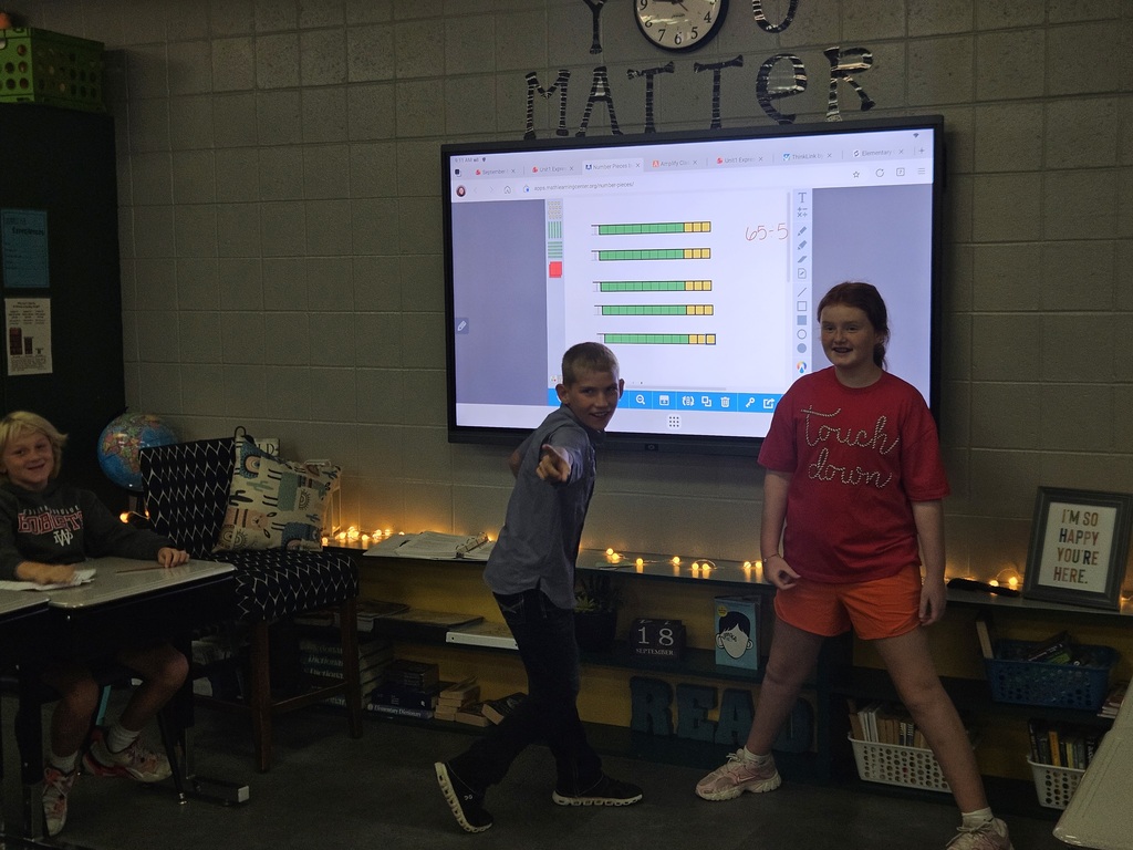 Two smiling students stand before an interactive display showing colorful math models during a classroom activity.