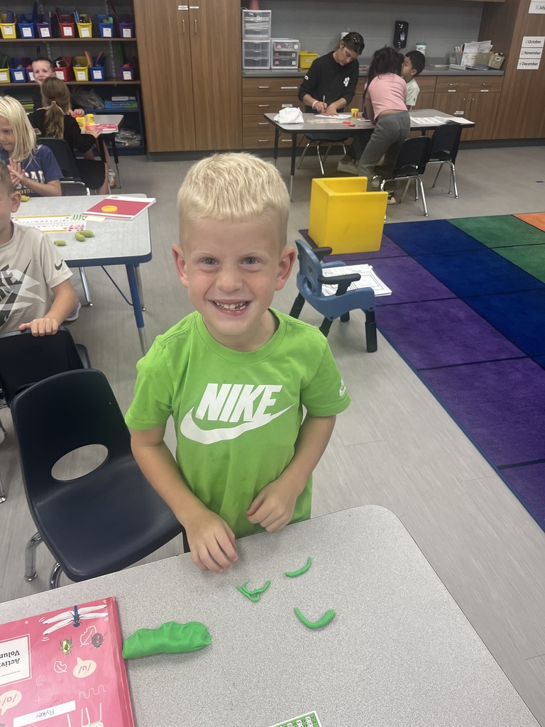 A smiling student stands at a classroom table with several green Play-Doh shapes arranged in front of him.
