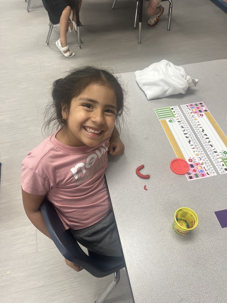 A student smiles beside a classroom table while creating curved shapes from red Play-Doh.