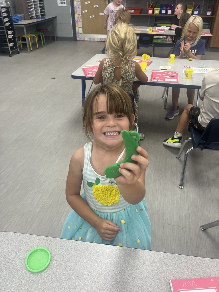 A smiling student proudly holds up a green Play-Doh creation while classmates work with modeling dough behind her.