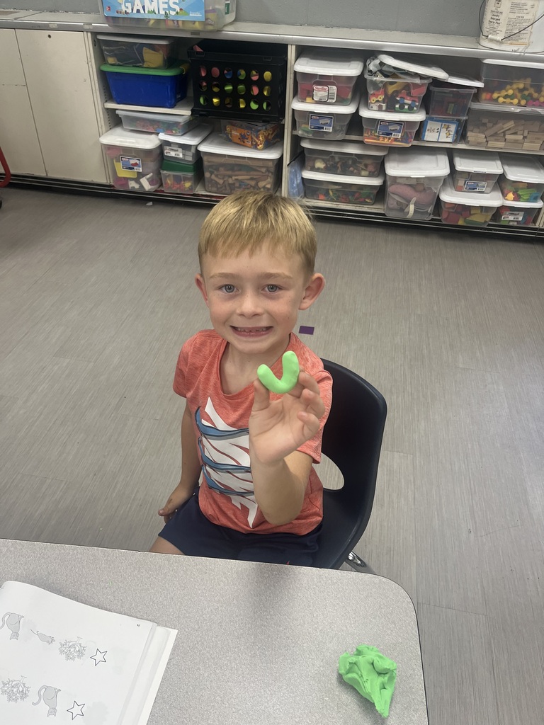 A smiling student holds up a curved green Play-Doh creation during a hands-on classroom activity.