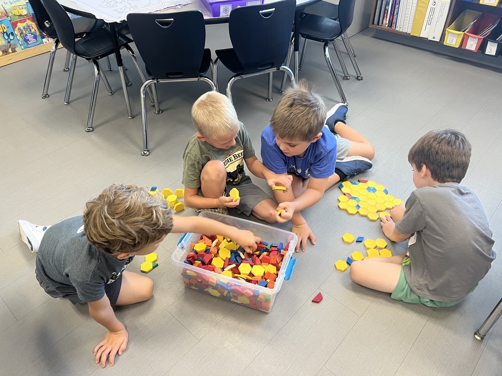 Four students sit on the classroom floor, sorting colorful geometric tiles and arranging yellow hexagons into patterns.