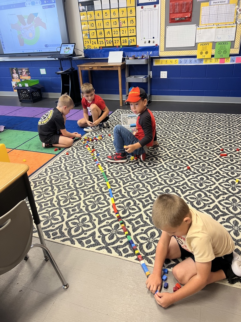 Four students work together on a classroom rug, creating a long, colorful line of connecting math cubes.