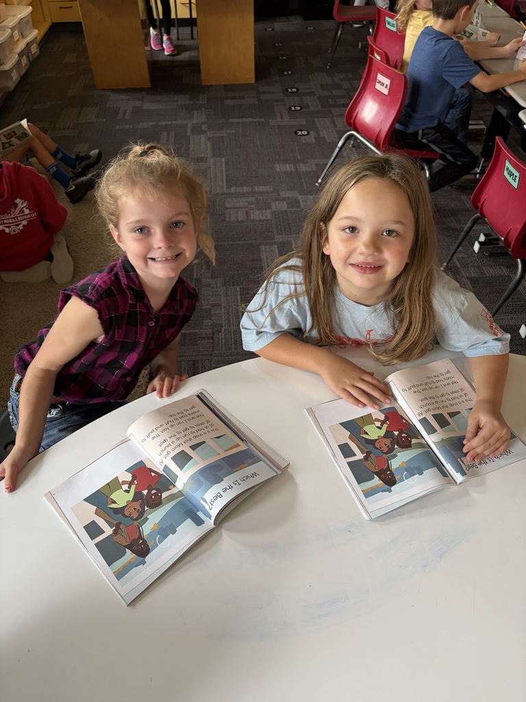 Two smiling students sit together at a table with matching open books during partner reading time.
