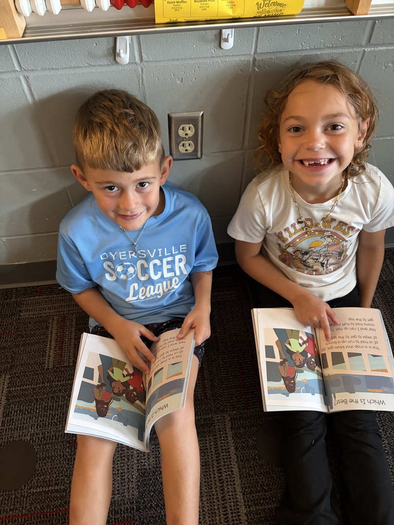 Two students smile at the camera while seated on the classroom floor with open books in their laps.