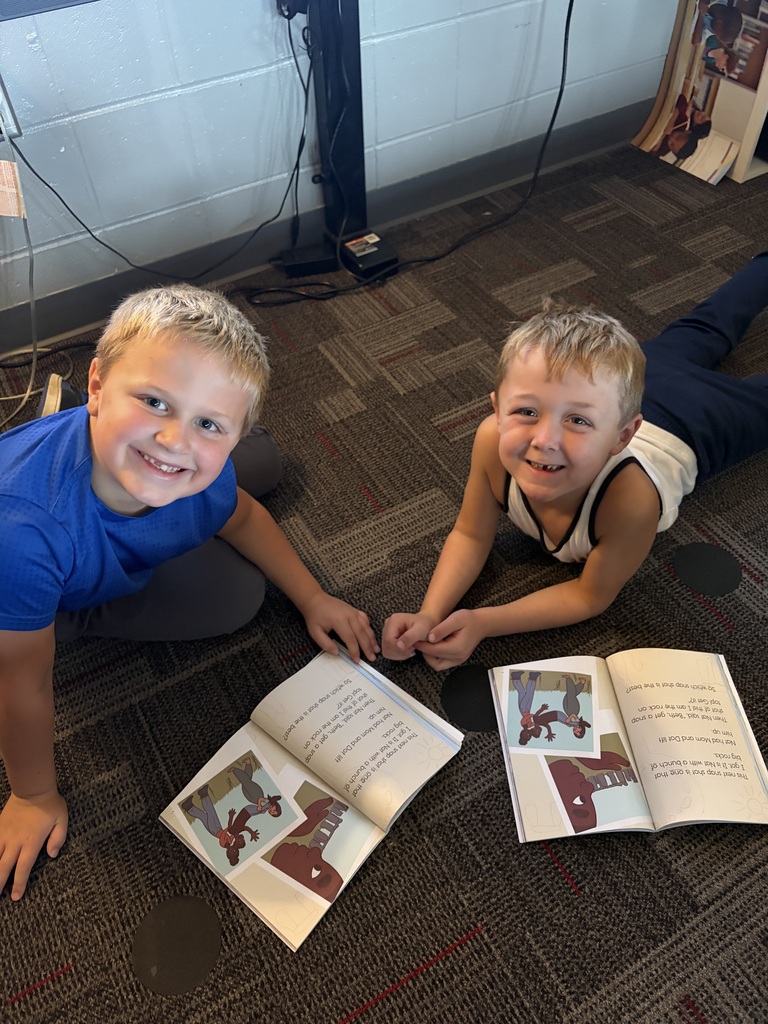 Two smiling students lie and sit on the classroom floor beside open books during partner reading time.