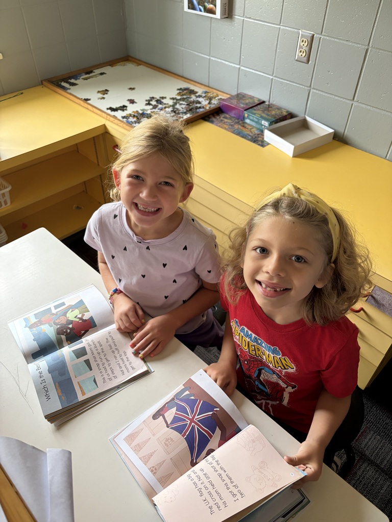 Two smiling students sit at a classroom table with open books during a partner reading activity.
