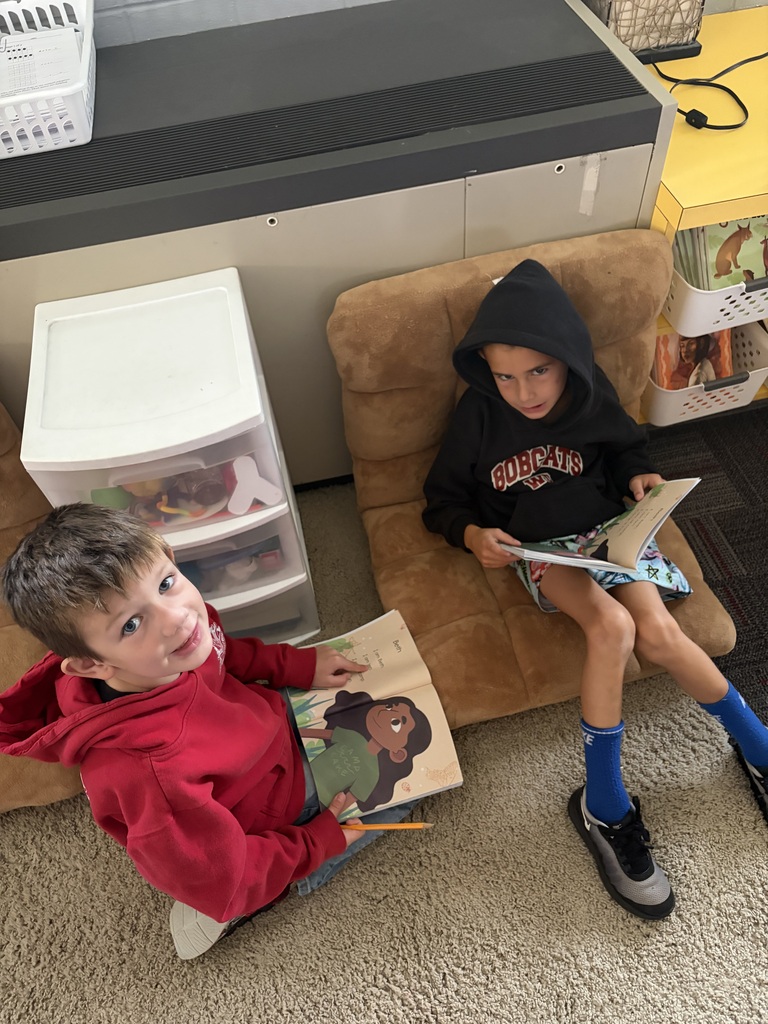 Two students read books in a cozy classroom reading area, one seated on the floor and one in a cushioned chair.