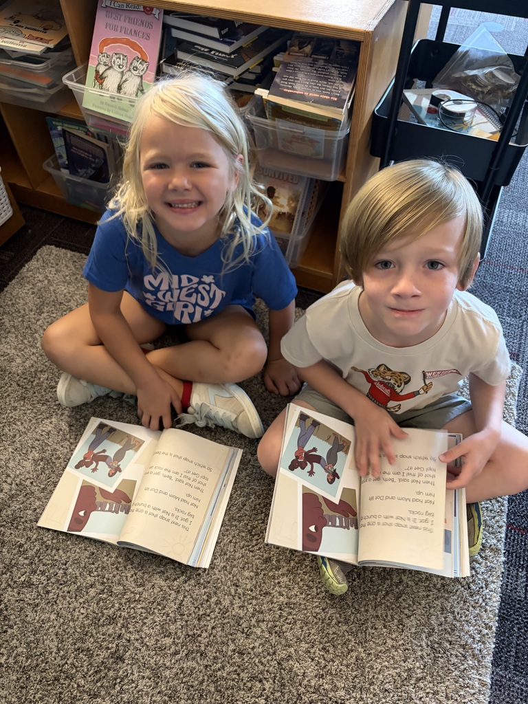 Two students sit together on a classroom rug, smiling with open illustrated books in front of them.