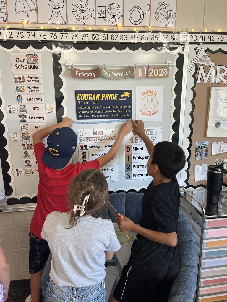 Three students work together to measure a classroom bulletin board with a ruler during a hands-on learning activity.