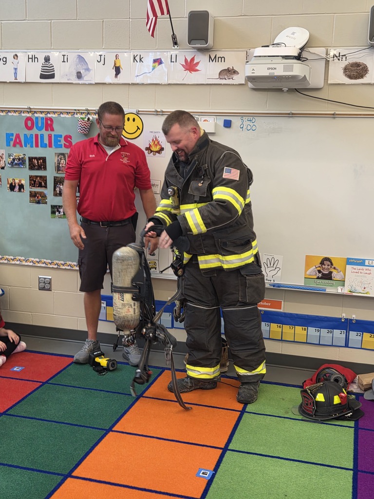 Firefighter demonstrates an air tank and breathing apparatus while another presenter watches in a kindergarten classroom.