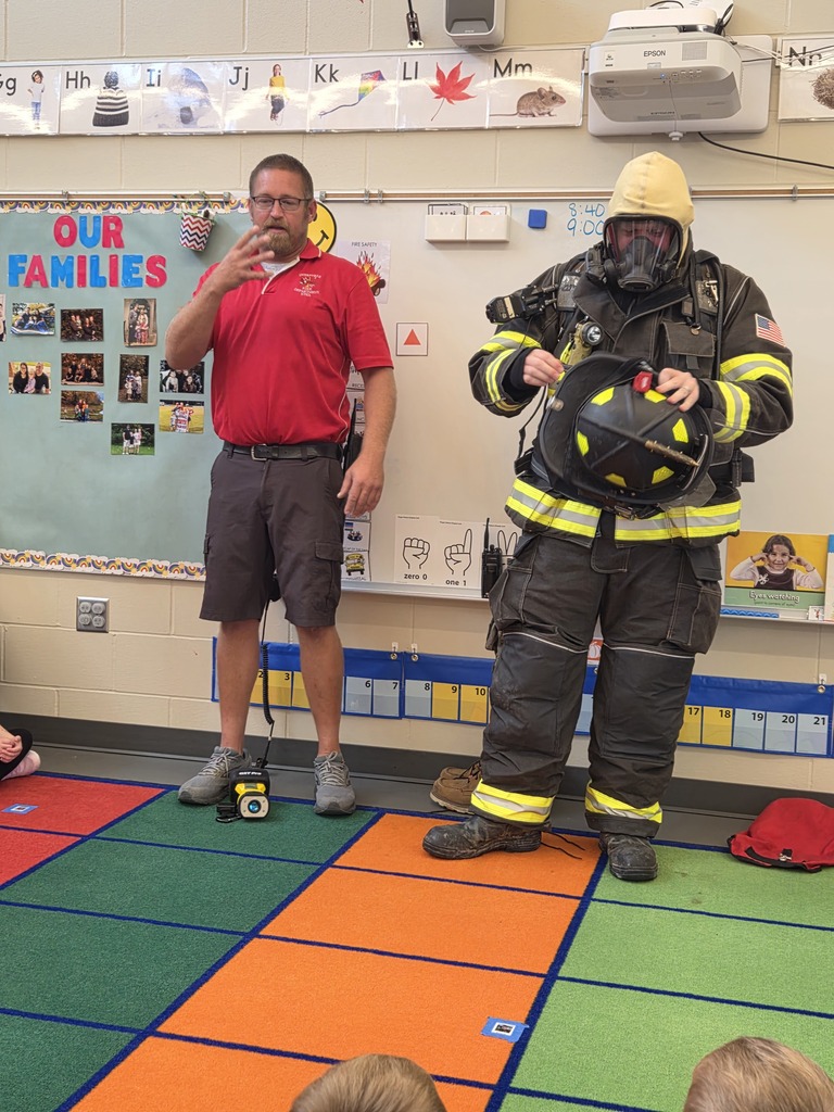Firefighter wearing a breathing mask holds his helmet while another presenter explains fire safety to young students.