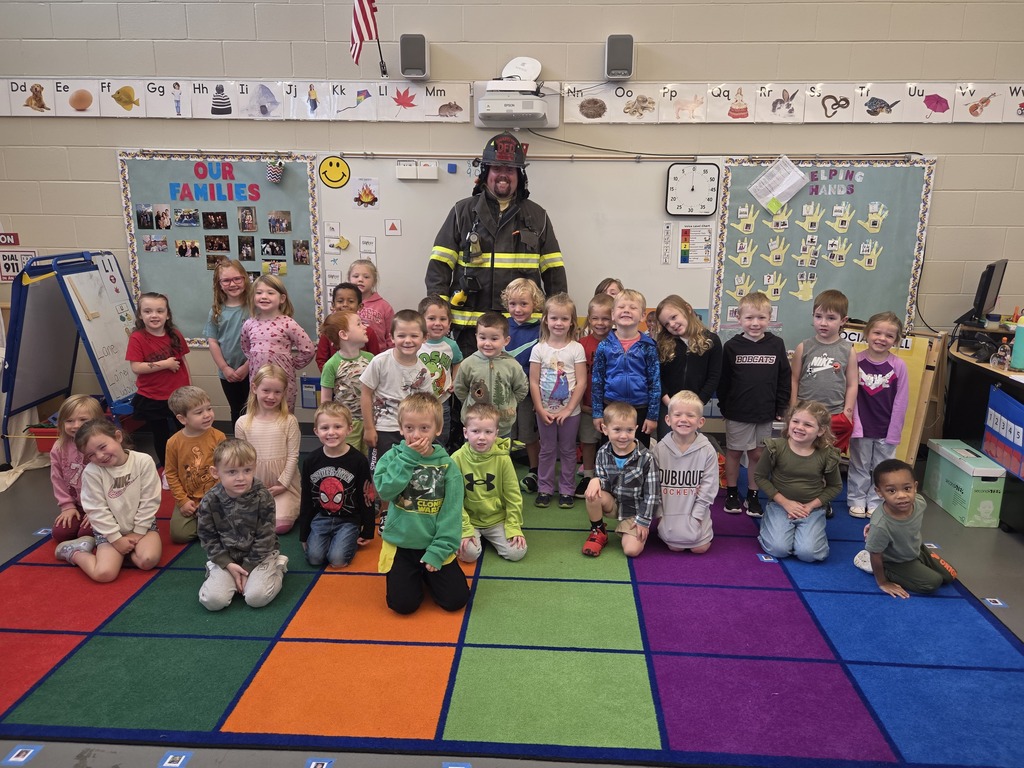 Kindergarten students pose for a group photo with a firefighter in full protective gear on a colorful classroom rug.