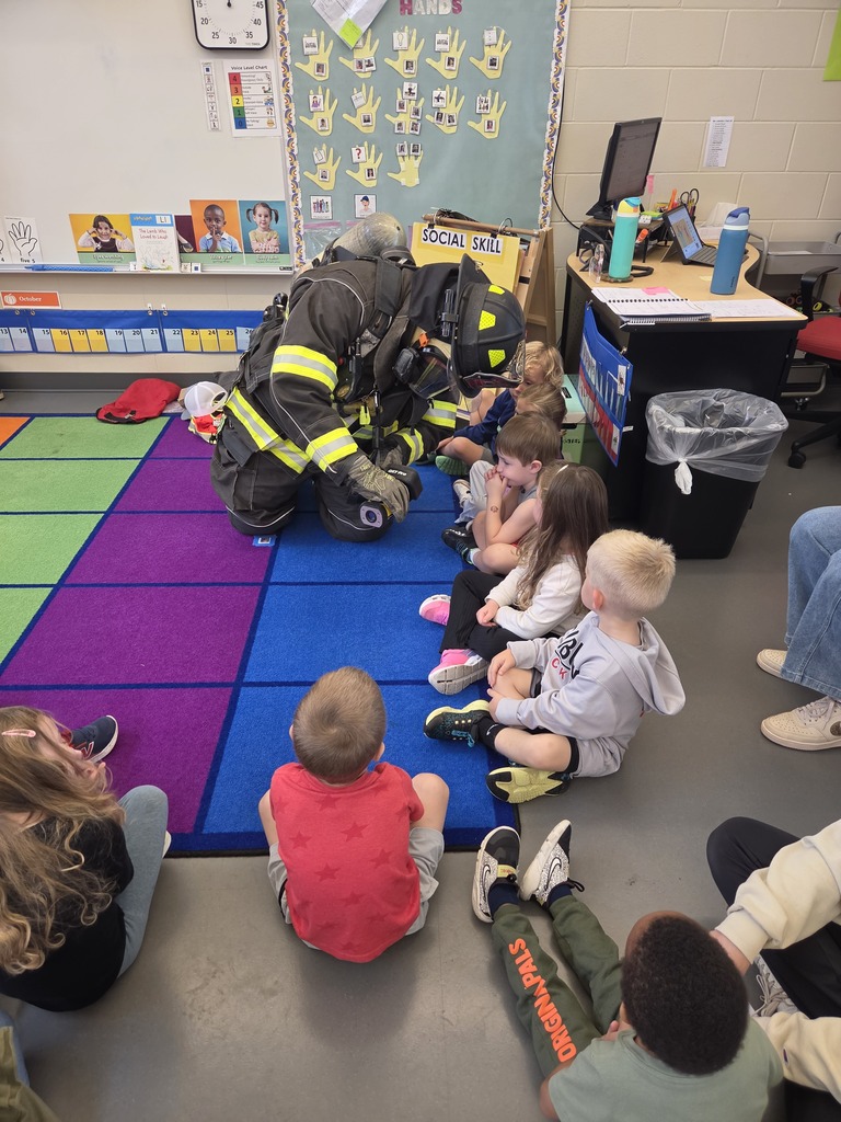 Firefighter in full protective gear kneels on a colorful classroom rug, showing equipment to a group of young students.