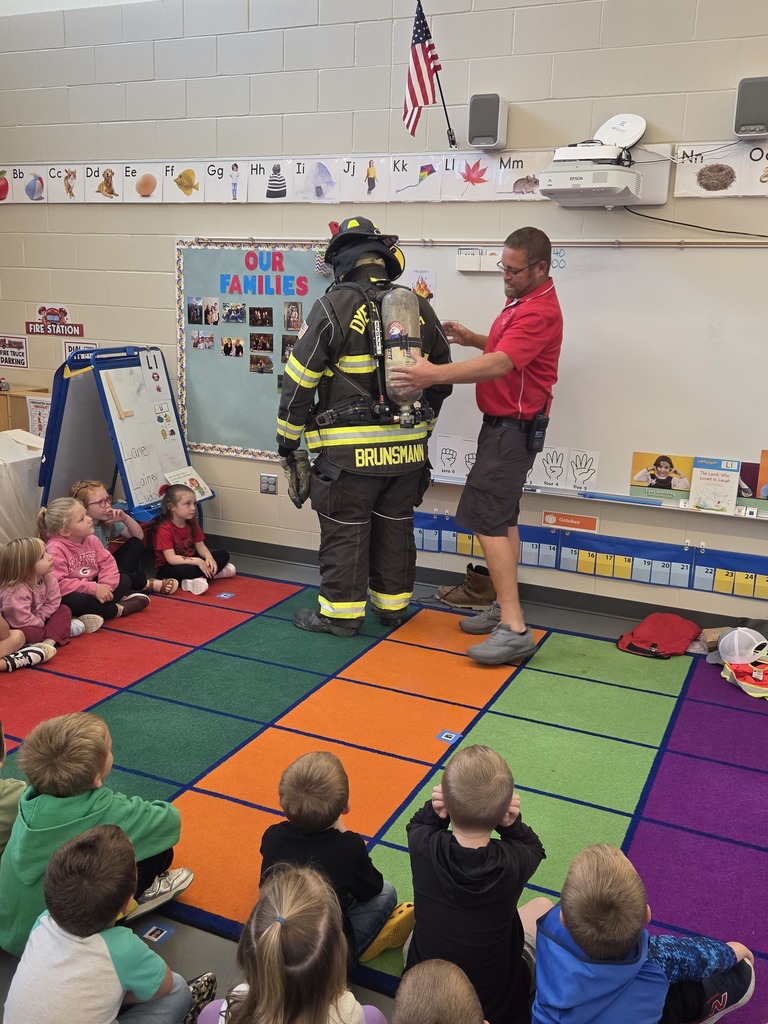 Firefighter wearing an air tank stands as a presenter adjusts the equipment while seated students watch the demonstration.