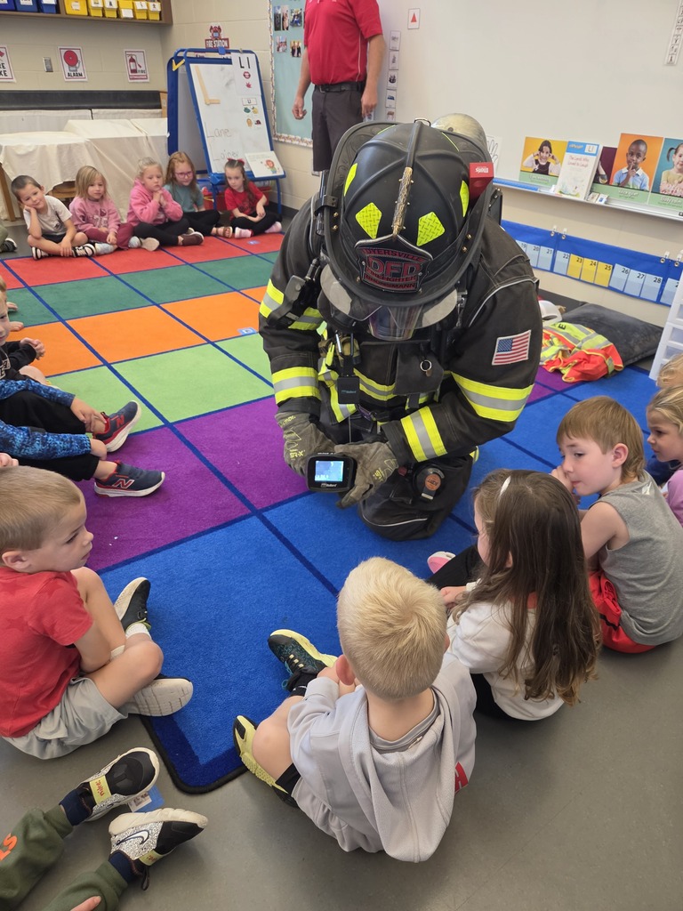 Firefighter kneels in full protective gear, showing a thermal imaging camera to kindergarten students seated around him.