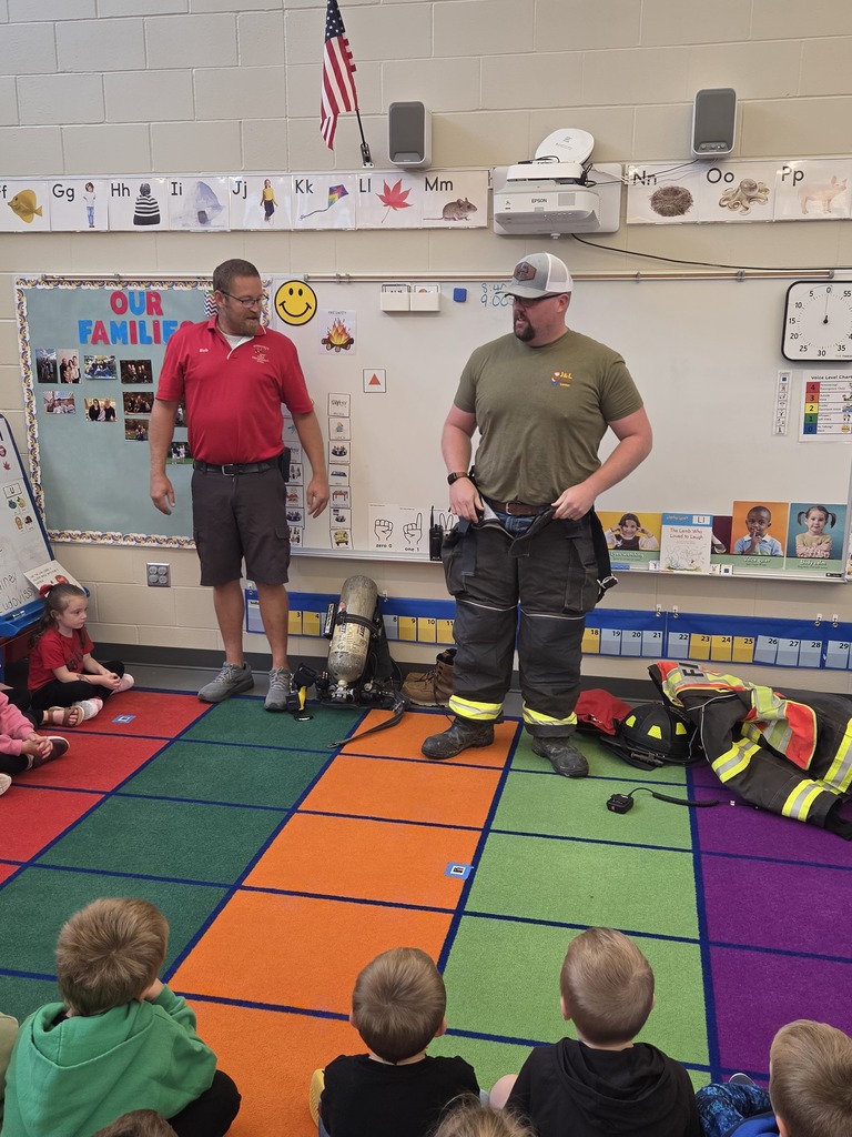 Two firefighters discuss protective clothing and safety equipment with kindergarten students seated on a classroom rug.
