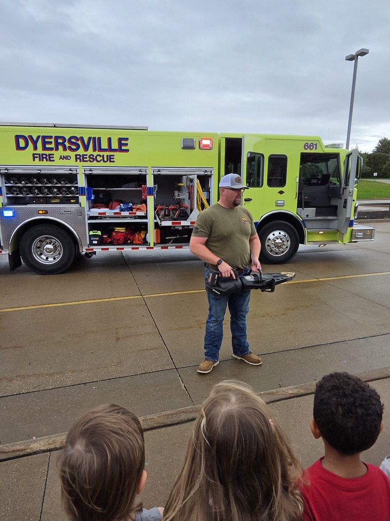 Firefighter demonstrates rescue equipment to students outside a bright yellow Dyersville Fire and Rescue truck.