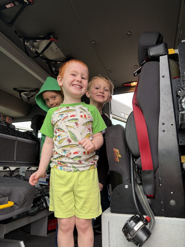 Three smiling children explore the inside of a fire truck, standing beside the driver's seat and emergency equipment.