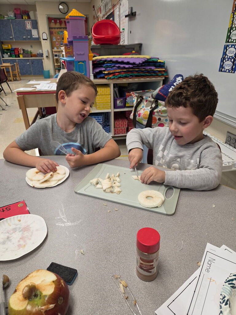 Two young students prepare apple slices at a classroom table, practicing cutting skills with plastic knives.