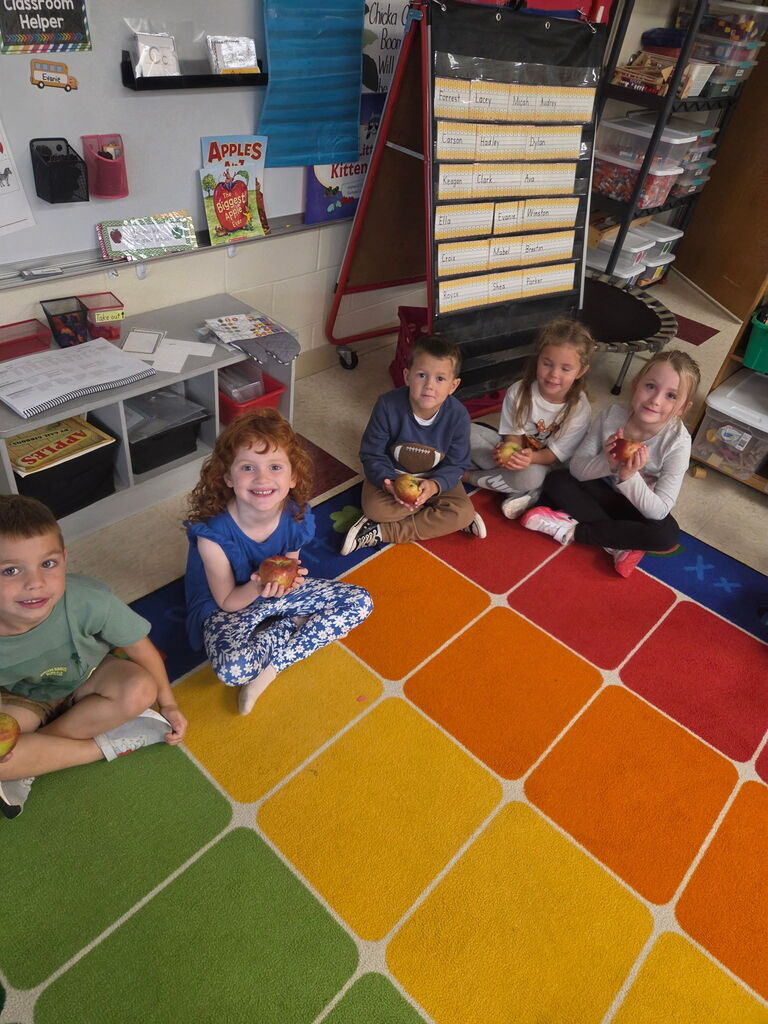 Five smiling students sit on a colorful classroom rug, holding fresh apples during a hands-on learning activity.