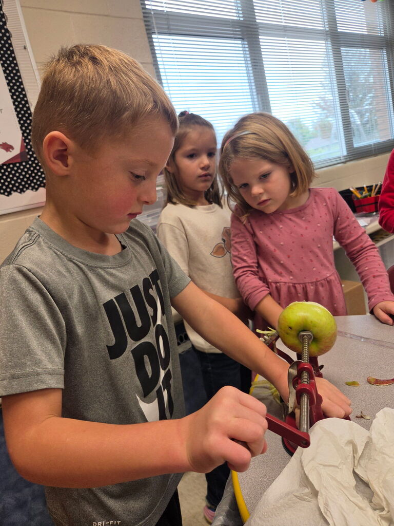A young boy turns the handle of an apple peeler while two classmates watch the apple preparation activity.