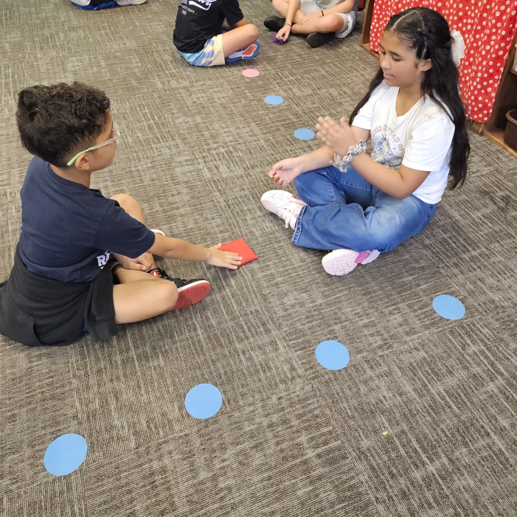 A boy and girl sit facing each other, using hand gestures and a red beanbag during a classroom partner activity.