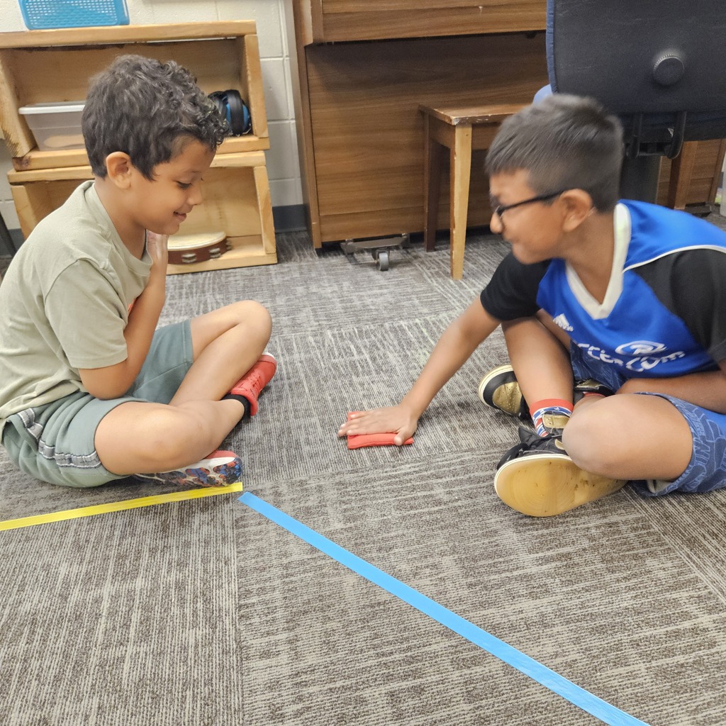 Two boys smile while sitting across from each other, playing a hands-on game with a red beanbag.