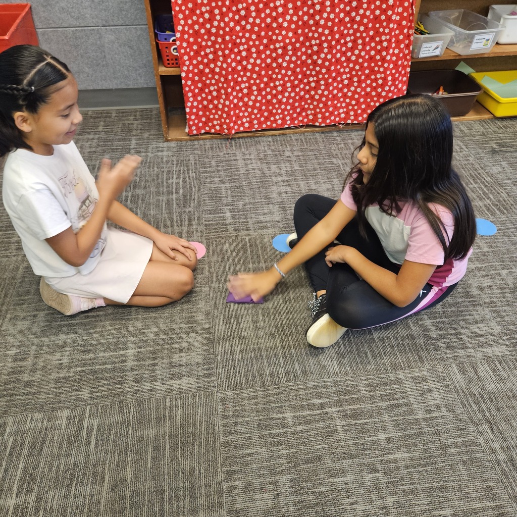 Two girls sit across from each other on the carpet, smiling and playing a hand-clapping game with a purple beanbag.