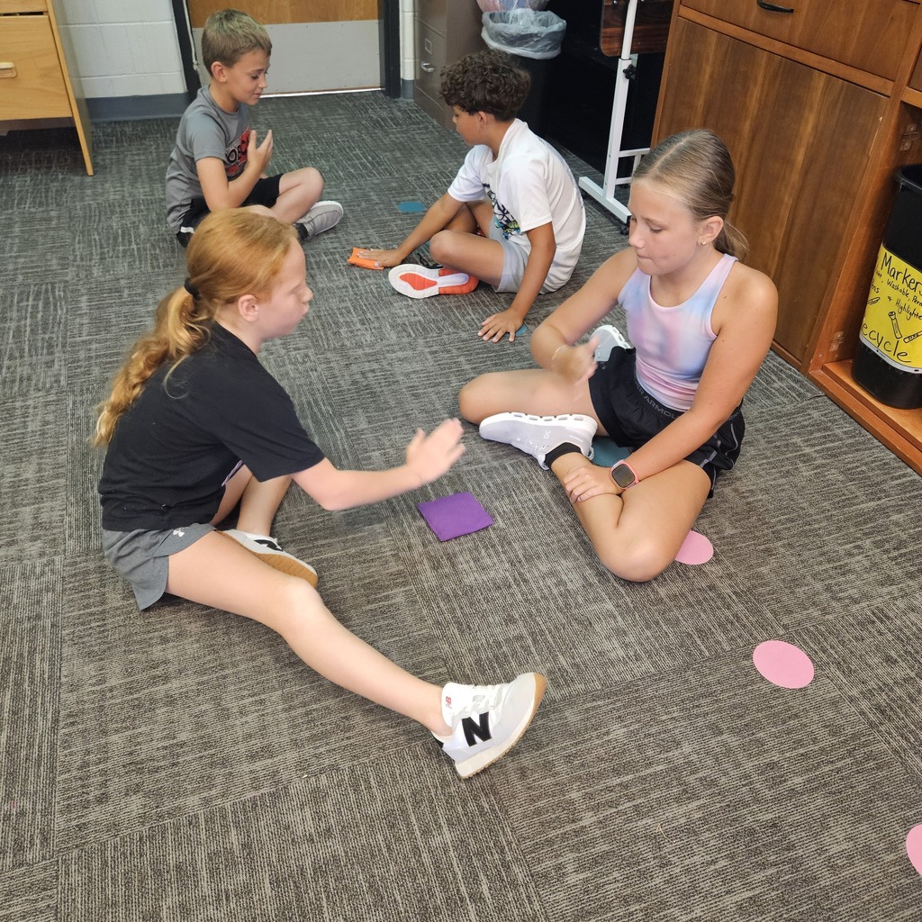 Four students work in pairs on a classroom carpet, practicing hand gestures and games with colorful beanbags.