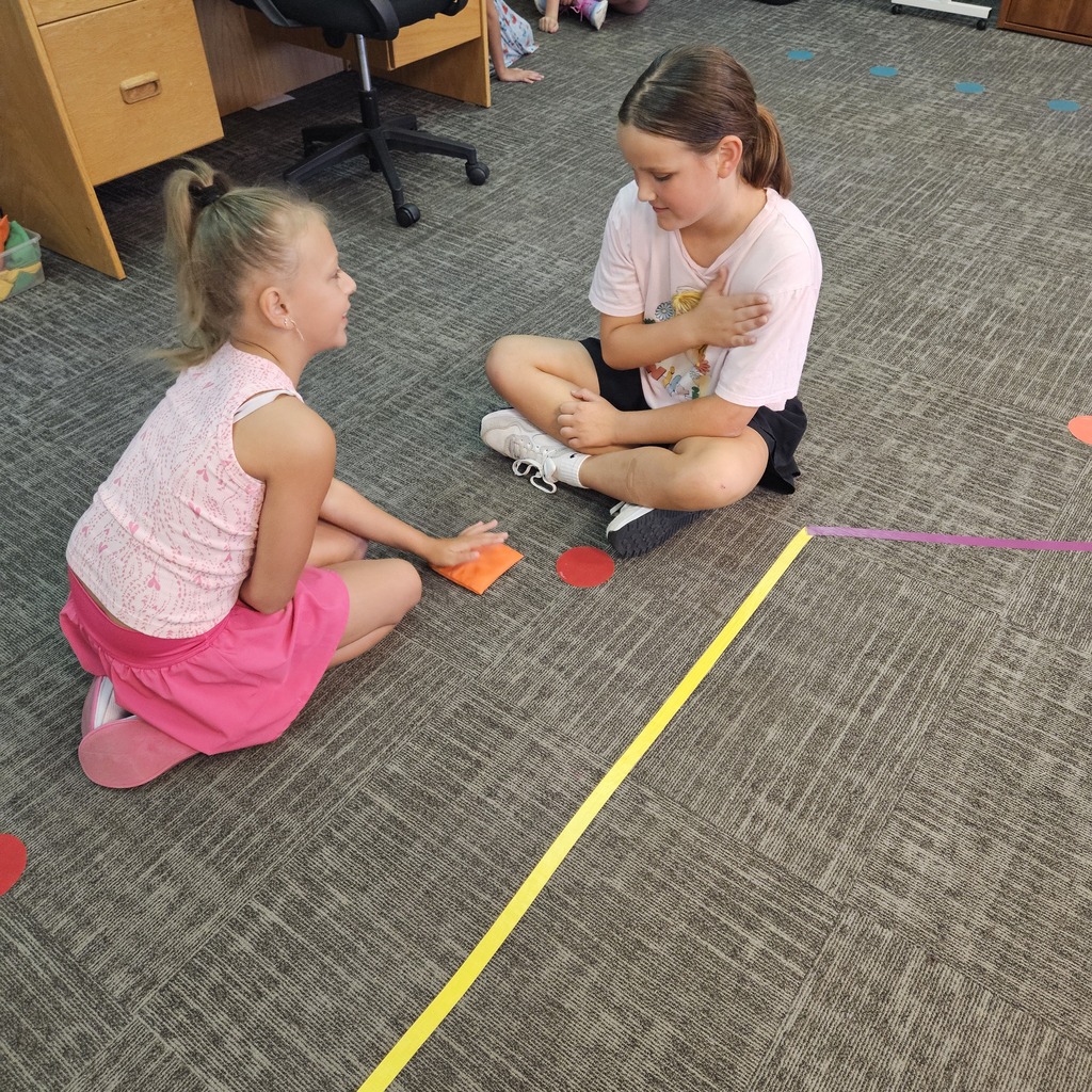 Two girls sit facing each other on a classroom carpet, using hand gestures during a partner activity.