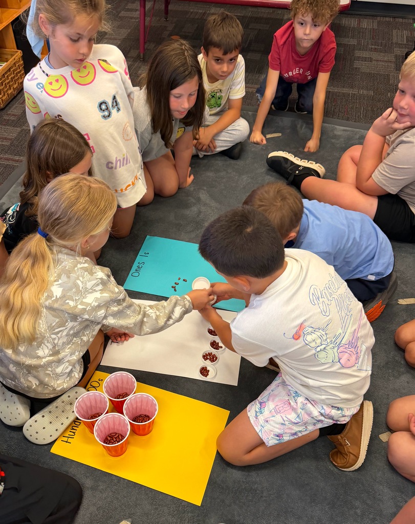 Students gather on a classroom rug to sort beans into ones, tens, and hundreds during a hands-on place value activity.