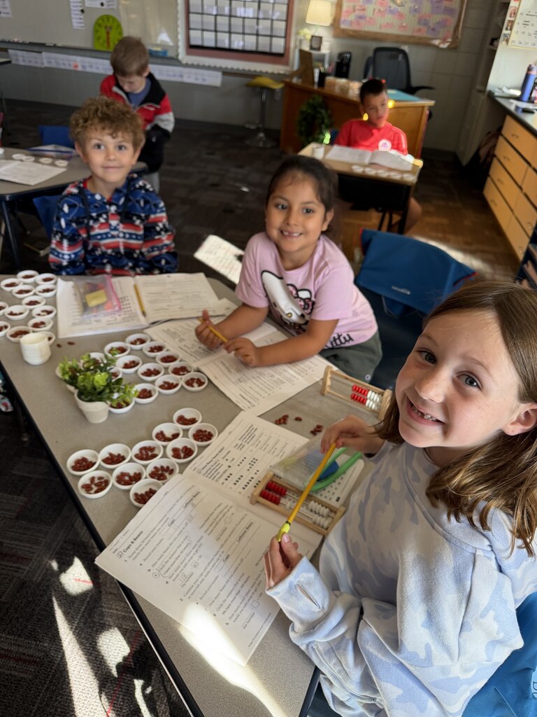 Smiling students practice math using beans, counting cups, abacuses, and worksheets at classroom desks.