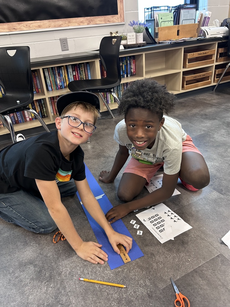 Two boys smile while measuring and arranging fraction pieces on a blue paper strip during a classroom math activity.